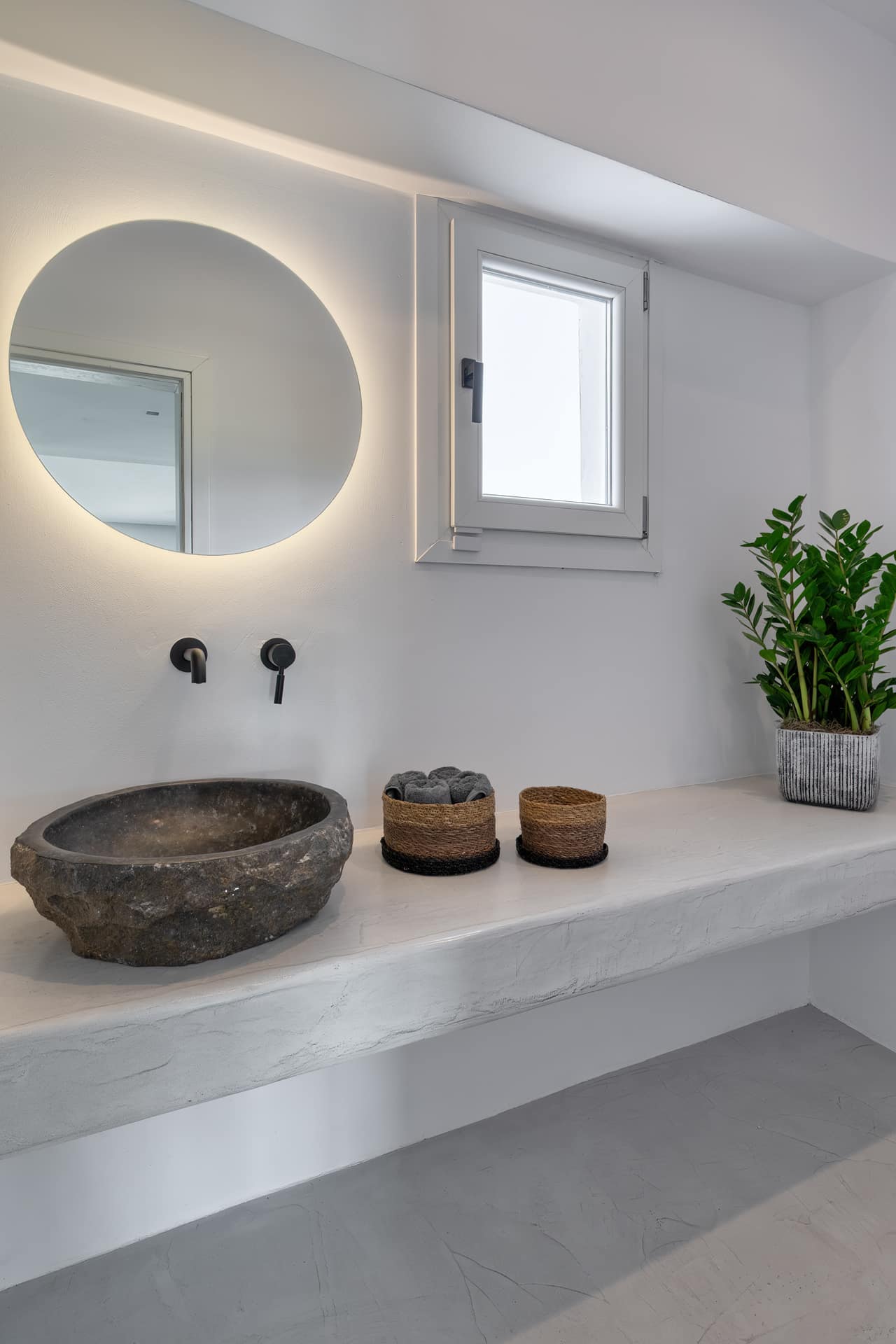 A raw stone basin on a microcement shelf beneath a round backlit mirror