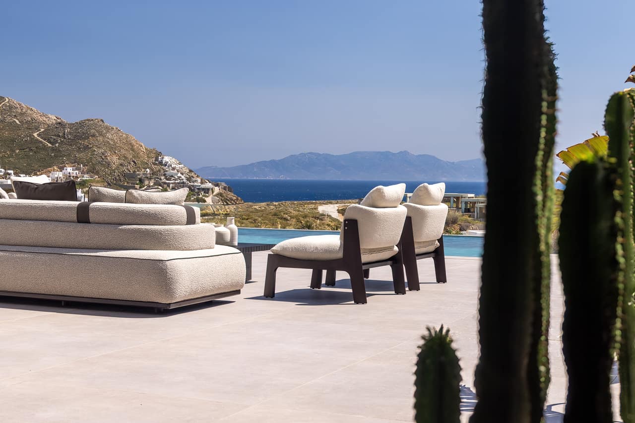 The terrace in clear daylight — bouclé seating, the pool, the Aegean beyond